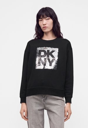 Jeune personne aux longs cheveux foncés portant un sweat-shirt noir avec le logo "DKNY" en sequins argentés et un jean gris clair sur un fond uni.