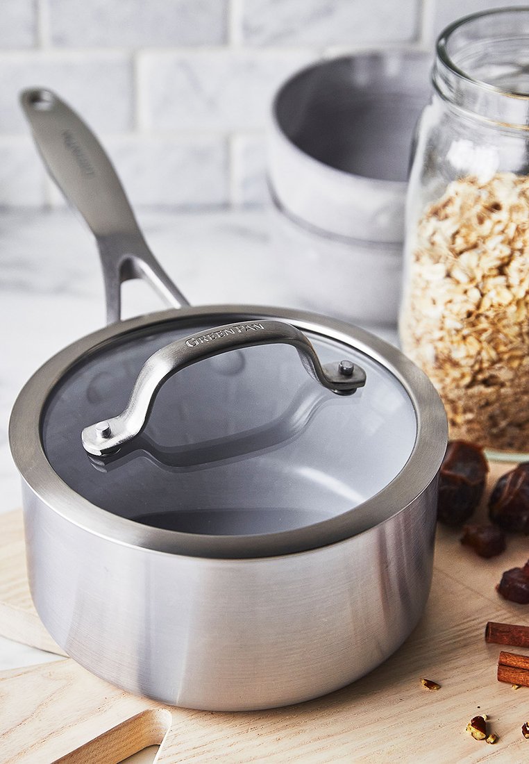 Edelstahl-Topf mit einem klaren Glasdeckel, ausgestattet mit einem silbernen Griff und einer polierten Oberfläche. Auf einer Holzoberfläche platziert, mit Zutaten in der Nähe.