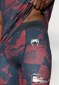 Μπλε και βουργουνδί κολάν με υφασματόμορφο σχέδιο, με μαύρη ελαστική μέση και λευκή λεπτομέρεια λογότυπου στο μηρό.