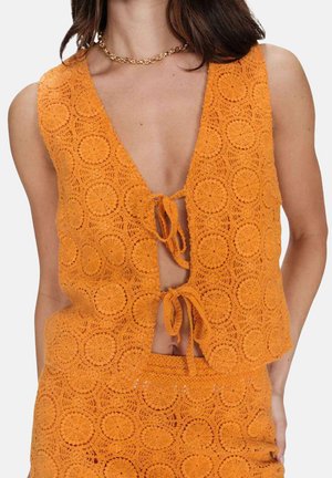 Femme portant un gilet en crochet orange sans manches avec des motifs circulaires, noué avec deux nœuds à l'avant, assorti d'une jupe coordonnée.