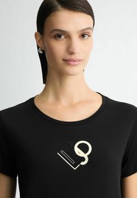 Zwarte T-shirt met een ronde halslijn, voorzien van een gouden grafisch ontwerp op de borst, benadrukt door kleine zwarte details. Glad katoenen materiaal.
