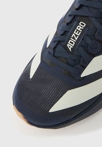 Zapatilla deportiva azul con parte superior de tela texturizada, detalles blancos, puntera redondeada y suela de goma color marrón claro. Cuenta con cordones y etiqueta con la marca.