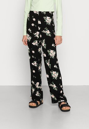 Kvinde iført sorte bukser med vide ben med hvidt og pink blomsterprint, lysegrøn langærmet top og sorte sandaler med korksåler.
