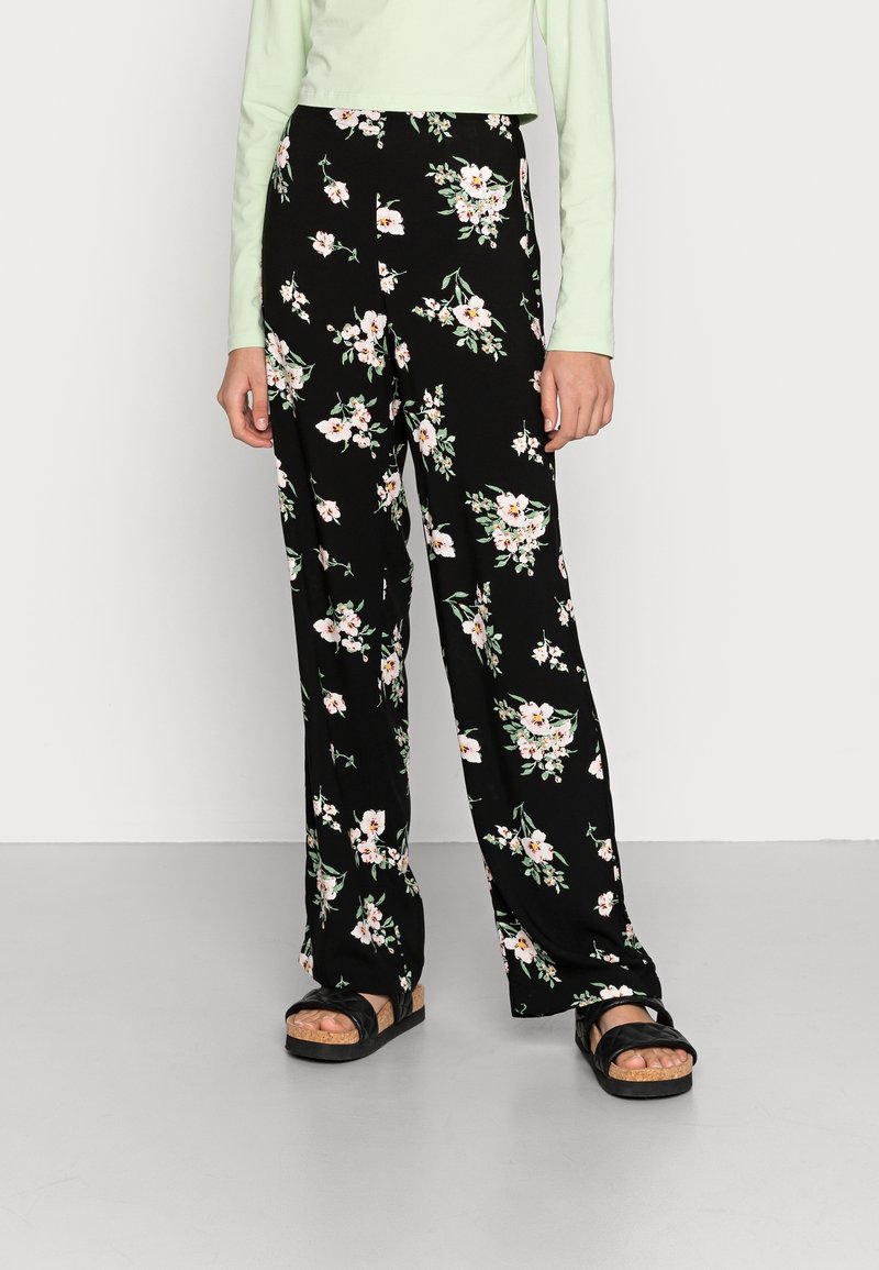 Femme portant un pantalon noir à jambes larges avec imprimé floral blanc et rose, un haut manches longues vert clair et des sandales noires à semelles en liège.