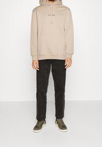 Felpa beige con cappuccio e tasca frontale, decorata con testo nero, abbinata a pantaloni in velluto a coste scuri e scarpe da ginnastica verde oliva con dettagli bianchi.
