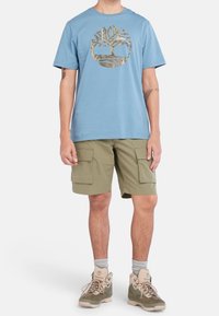 Osoba v modrém tričku s logem stromu, zelených cargo šortkách, šedých ponožkách a zelených turistických botách stojící na bílém pozadí.