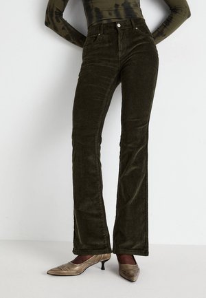 Femme portant un pantalon flare en velours côtelé taille haute de couleur olive foncé avec des chaussures beige à talons bas, debout devant un fond clair uni.