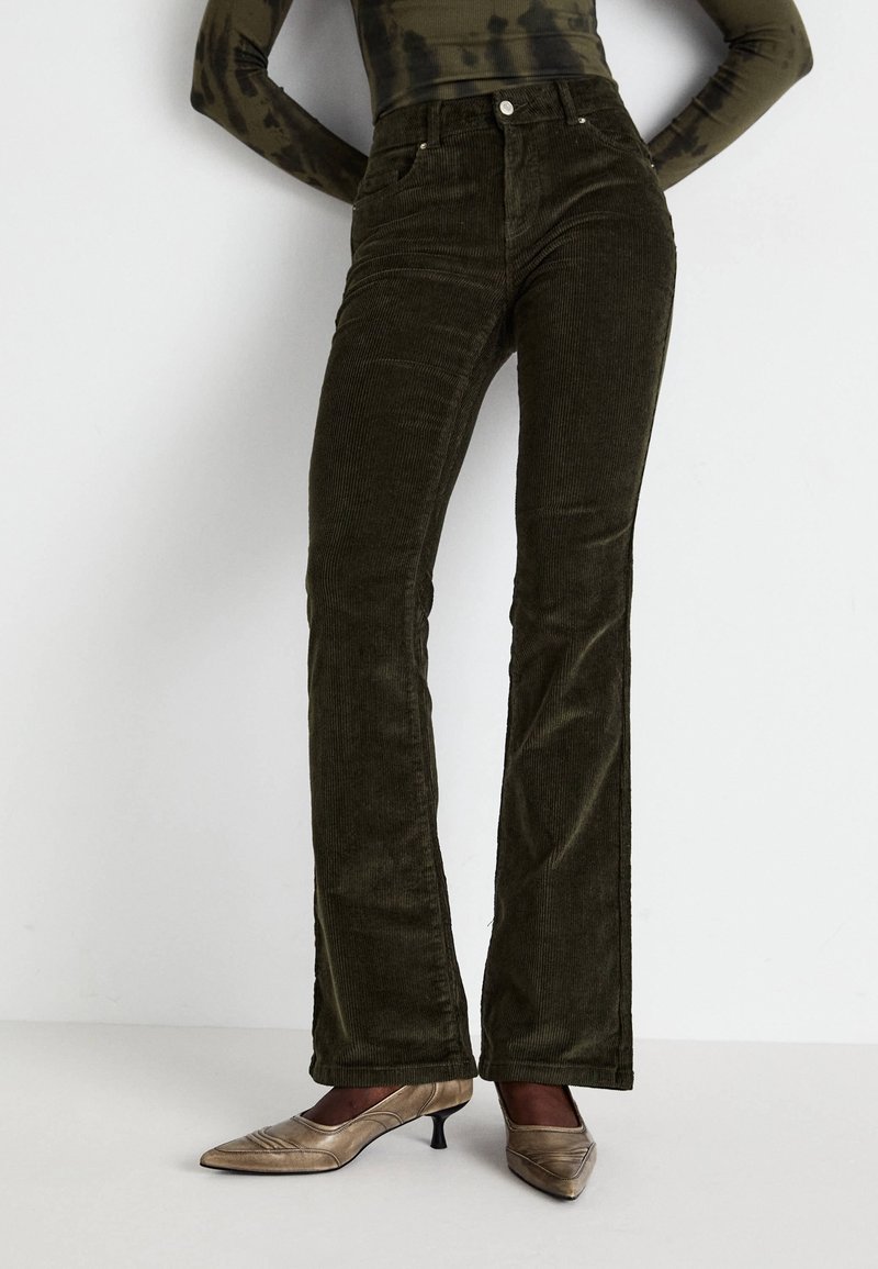 Femme portant un pantalon flare en velours côtelé taille haute de couleur olive foncé avec des chaussures beige à talons bas, debout devant un fond clair uni.