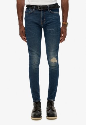 Jeans de denim oscuro de corte ajustado con parches desgastados y un ligero desvanecimiento; combinados con botas negras con cordones y un cinturón trenzado negro.
