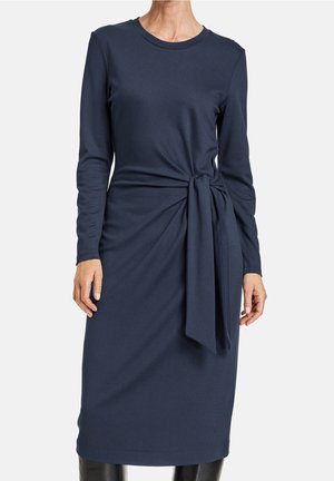 Granatowa sukienka o długości do kolan z długim rękawem, okrągłym dekoltem i wiązaniem w talii na stojącej osobie.