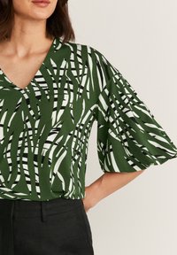 Donna che indossa una blusa a V con maniche corte larghe, astratta nei colori verde, bianco e nero, abbinata a pantaloni neri, su uno sfondo neutro.