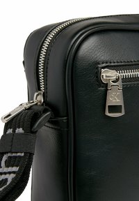 Borsa in pelle nera con una texture liscia, zip argentata e una tasca laterale con zip, impreziosita da un dettaglio metallico con marchio e una tracolla regolabile.