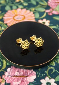 Orecchini d'oro a forma floreale con dettagli intagliati, adagiati su un piatto nero lucido, su uno sfondo floreale colorato.