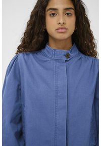 Modra bombažna jakna z visokim ovratnikom, zapiranjem na gumb in napihnjenimi rameni, ki se ponaša z gladko teksturo in čistimi oblikovalskimi linijami.