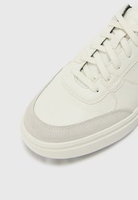 White leather sneaker with grey suede accents, rounded toe, textured surface, and lace-up design. Flat white sole with subtle stitching detail.