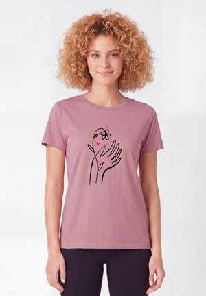 Frau mit lockigem Haar, die ein pinkes T-Shirt mit einem schwarzen Linienkunst-Design von zwei Händen, einer Blume und einem kleinen roten Herz auf der Brust trägt.