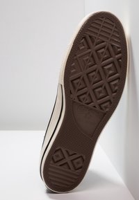 Schwarze Gummisohle eines Sneakers mit einer strukturierten braunen Außensohle, die ein geometrisches Muster aufweist. Ein beiger Rand verläuft seitlich entlang für mehr Stabilität.