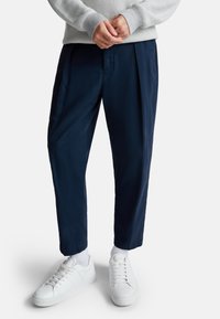 Marineblauwe broek met een ontspannen pasvorm en plooien, gecombineerd met witte sneakers en sokken. De broek heeft een gladde textuur en is ontworpen met een taps toelopende pijp.