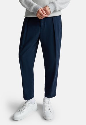Navy trousers with a relaxed fit and pleats, paired with white sneakers and socks. Trousers feature a smooth texture and tapered leg design.