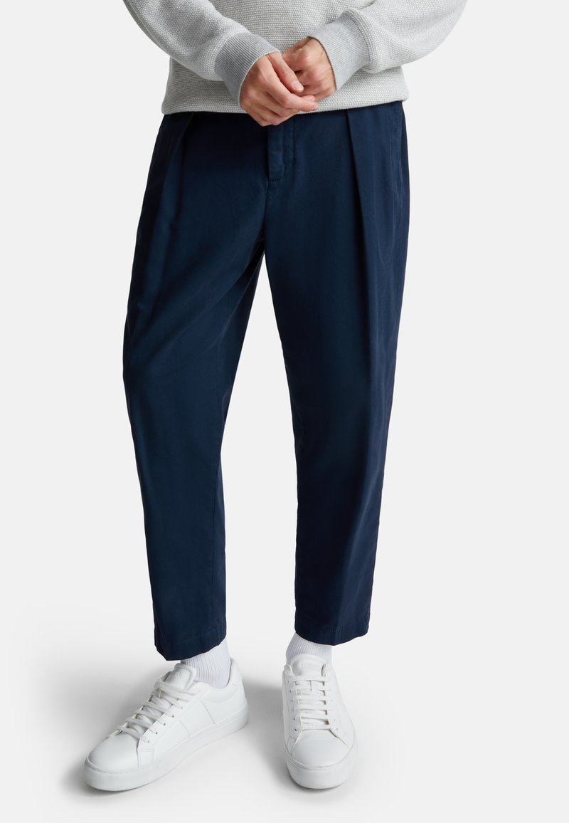 Marineblauwe broek met een ontspannen pasvorm en plooien, gecombineerd met witte sneakers en sokken. De broek heeft een gladde textuur en is ontworpen met een taps toelopende pijp.