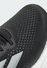 Black athletic shoe with a textured fabric upper, featuring a silver stripe logo and contrasting white sole; detailed lacing system.