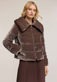 Giacca puffer in velluto marrone con colletto ampio, chiusura con zip e motivo trapuntato orizzontale. Maniche lunghe fino alle spalle e tasche laterali.