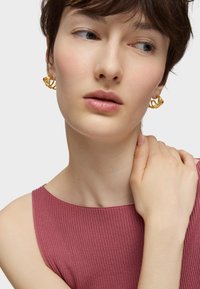 Boucles d'oreilles en or en forme de cercle avec un design entrelacé et un accent rouge, associées à un haut rose ajusté à côtes. Teint de peau lisse, arrière-plan neutre.