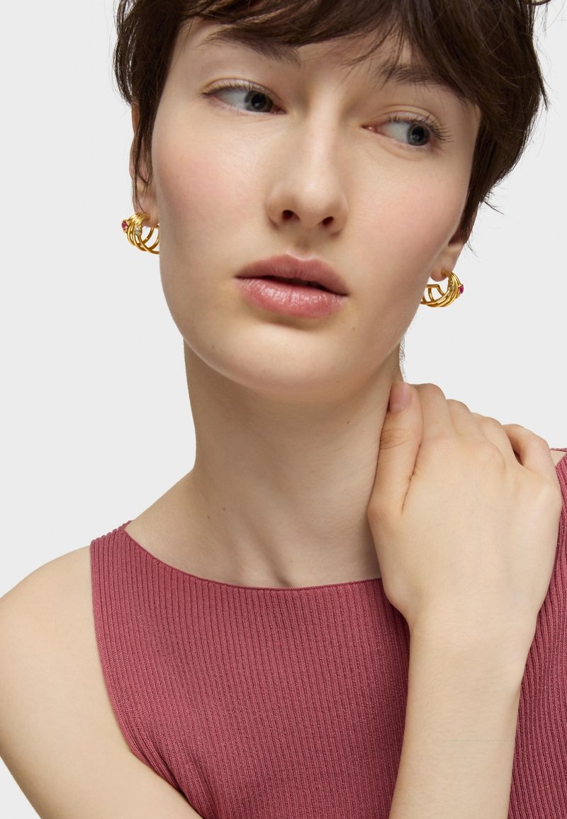 Boucles d'oreilles en or en forme de cercle avec un design entrelacé et un accent rouge, associées à un haut rose ajusté à côtes. Teint de peau lisse, arrière-plan neutre.