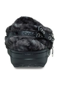 Black Crocs with a fluffy gray faux fur upper, featuring a black strap with decorative silver grommets and a small charm on the side.