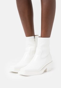 Botines blancos con acabado brillante, punta cuadrada, cremallera lateral y tacón ancho, que presentan un diseño moderno y elegante.