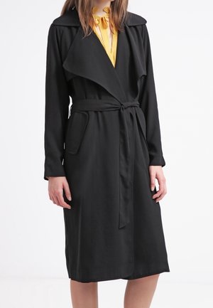 Frau mit schwarzem, gegürteten Trenchcoat über einem gelben Oberteil mit Schleifendetails, steht vor weißem Hintergrund.