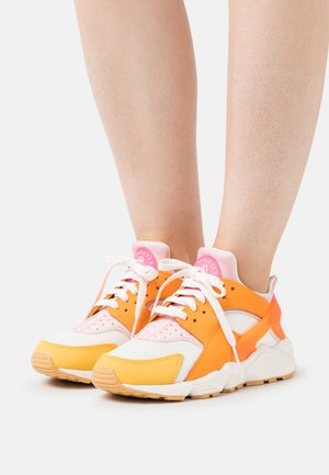 Scarpe sportive colorate con pannelli arancioni, rosa e bianchi, tessuto in rete, perforazioni e una suola in gomma spessa. I lacci sono bianchi.