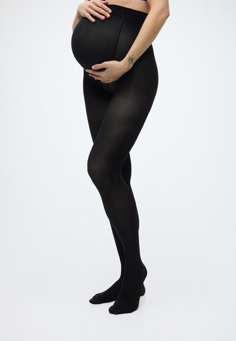 Έγκυος γυναίκα με μαύρο κολάν εγκυμοσύνης, τα χέρια της απαλά να στηρίζουν την κοιλιά της, στέκεται μπροστά σε ανοιχτό, απλό φόντο.