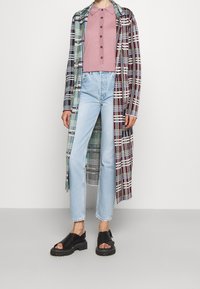 Abrigo largo de cuadros en burdeos, azul y verde, combinado con una camisa corta rosa con botones y jeans de color azul claro. Sandalias de plataforma negras.