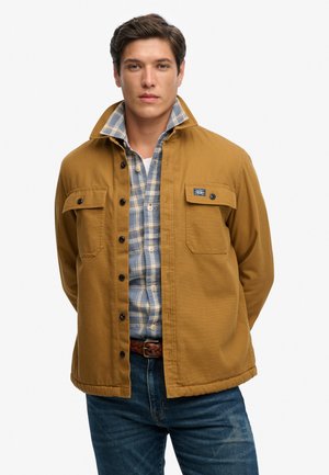 Mand iført en beige jakke med knapper over en blå- og hvidternet skjorte og blå jeans, stående med hænderne bag ryggen.