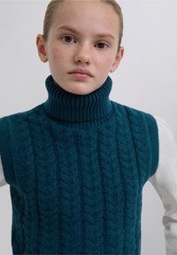 Türkisfarbene gestrickte Pulloverweste mit einem gerippten Rollkragen, die ein strukturiertes Zopfmuster und ein ärmelloses Design aufweist und über einem weißen Hemd getragen wird.