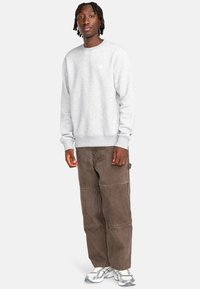 Felpa grigia con polsini a coste, abbinata a pantaloni cargo marroni con texture a patchwork. Il modello indossa scarpe da ginnastica argentate.
