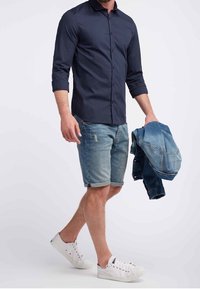 Marineblauwe button-up shirt, lichtblauwe denimshorts, witte sneakers en een blauwe denimjasje in de hand. Heeft een casual, relaxte pasvorm.