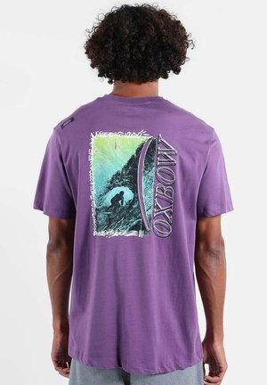Un homme aux cheveux bouclés porte un t-shirt violet affichant un surfeur chevauchant une vague et le mot "OXBOW" au dos.