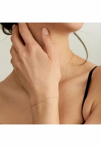 Goldkette-Armband mit kleinen, unregelmäßig verteilten, strukturierten Akzenten, getragen an einer Hand in der Nähe eines Halses, der mit mehreren Goldketten geschmückt ist.