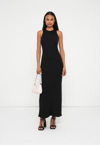 Rochie maxi neagră, fără mâneci, cu o siluetă dreaptă, având embellishments subtile. Modelul ține o geantă mică de culoare roz pal și poartă tocuri negre.