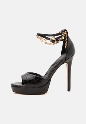 Sandale noire à talons hauts avec semelle plateforme, texture croco, bride de cheville et détail chaîne dorée sur un fond clair uni.