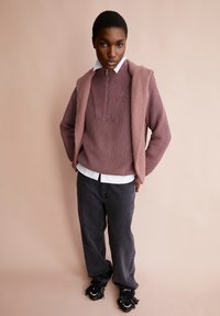 Gebreide pullover in gedempt mauve met een geribbelde textuur, met een ritssluiting aan de hals, gedragen over een witte blouse met kraag. Brede donkere spijkerbroek en zwarte fringeschoenen maken de look compleet.