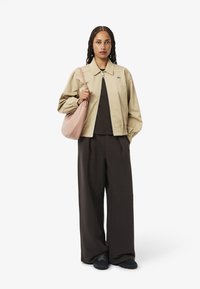 Giacca bomber beige con polsini elastici, top nero, pantaloni larghi marroni, e scarpe nere; tiene una borsa rosa chiaro. Texture liscia, vestibilità rilassata.