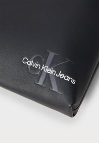 Melnās mākslīgās ādas maiss, kas iezīmēts ar izspiedu Calvin Klein Jeans logotipu, ar gludu tekstūru, noapaļotām malām un smalku spīdumu.
