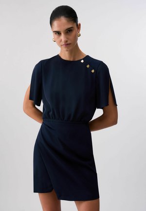 Kobieta ubrana w granatową sukienkę z rozcięciami na rękawach, złotymi guzikami na ramieniu i złotymi kolczykami kołami, stojąca na tle jednolitego jasnego tła.