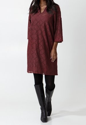 Kobieta ubrana w bordową koronkową sukienkę o długości do kolan z rękawami do łokci, czarne rajstopy i czarne kozaki za kolano, stojąca na białym tle.