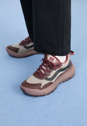 Par af bordeaux og beige Vans-sneakers med tykke såler båret med sorte bukser og hvide sokker på lyseblåt gulv.