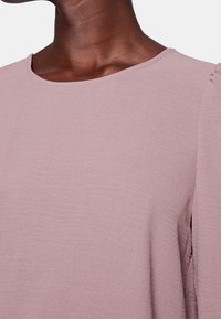 Haut rose clair texturé avec un col rond et un léger froncé subtil sur les manches, confectionné dans un tissu doux.