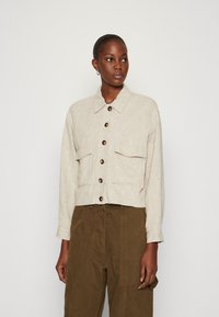 Beige linne cropped jack met een kraag, sluiting met knopen aan de voorkant, twee borstzakken en lange mouwen; gecombineerd met olijfgroene broeken.